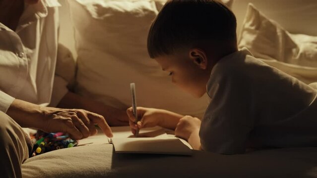 An elderly woman with dark hair in a white shirt sits with her grandson on a white sofa. Grandmother and grandson draw together with colored pencils. A cozy evening at Grandma's.