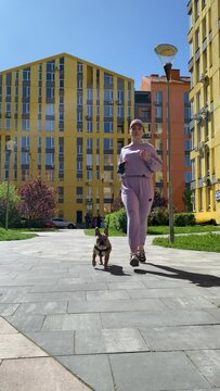 Vertical front view young adult woman in purple sportswear jogging dog walking at urban area sunny morning. Healthy female pet owner running domestic animal on leash outdoors. City lifestyle concept