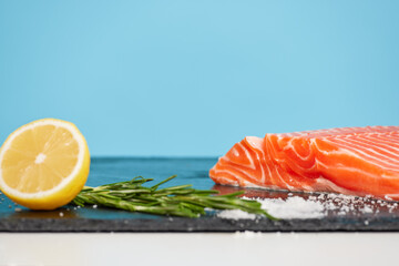 Slice of salmon with spices and lemon isolated on blue background, still-life with tasty fish fillet on black cutting board, seafood dish concept, copy space