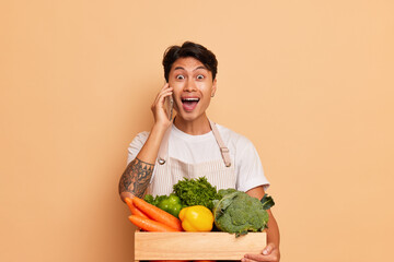 Cheerful young farmer stands with box of fresh vegetables, sells his vegetables, smiles joyfully while talking on the phone, organic food concept, copy space