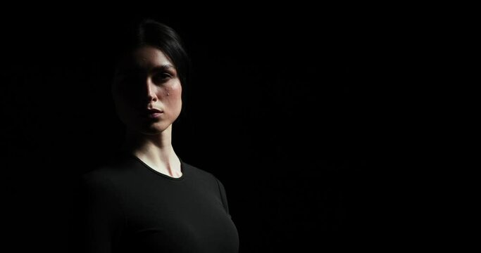 Woman stands alone on a black background, slowly and dramatically lifting her head and looking into the camera with a serious expression. She exudes loneliness and sadness.