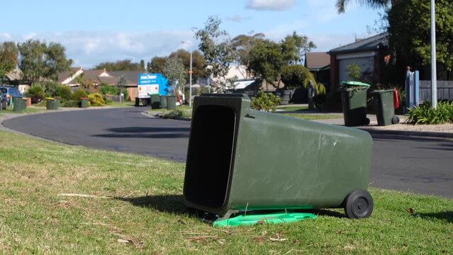 An empty green rubbish bin fell on the nature strip caused by a moving garbage truck collecting household waste on suburban street. Melbourne VIC Australia.