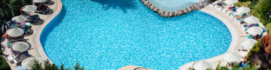 Chill out summer vacation in luxury resorts concept. Top view, a stunning blue curved swimming pool with sun loungers and umbrellas is surrounded by lush green tropical vegetation.