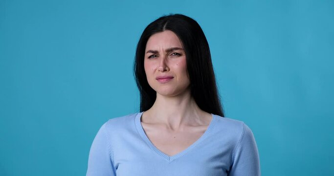 A woman of Caucasian nationality stands on a blue background, looking directly at the camera. Suddenly, she notices something unpleasant and leans back in slight disgust.