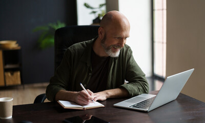 Smiling mid-aged man focused and productive state. He seated at desk indoors, engrossed work. With notepad in front, he diligently writing down notes, while simultaneously looking at computer screen. 