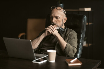 Dedicated elderly worker in loft office environment. With puzzled expression on his face, he sits attentively in front of a computer, deeply engaged in solving a complex problem. 