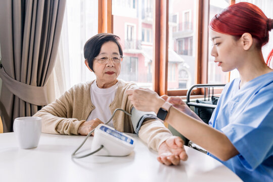 Nurse measuring blood pressure by using automatic blood pressure monitor on mature senior Asian woman with care. Caregiver visit at home. Home health care and nursing home concept.
