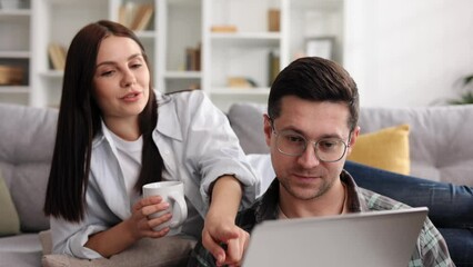 Cheerful young couple relaxing together with laptop and coffee in living room, happy young couple relaxing at home having hot drinks, using computer and shopping, close up shot. - Powered by Adobe