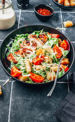 Homemade Caesar salad with crispy chicken and tomatoes on a black tiled background