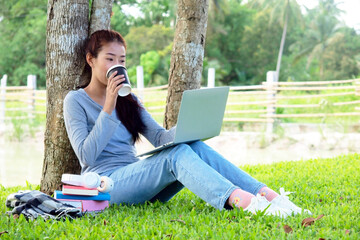 Asian student girl is studying  by laptop in nature with tree. Asian student is happy in study online and drinking coffee. Education in online life concept. Study Online in vacation.