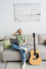 young woman with blonde and short hair, bangs and eyeglasses using laptop while sitting on comfortable couch and looking at camera near guitar in modern living room with painting on wall