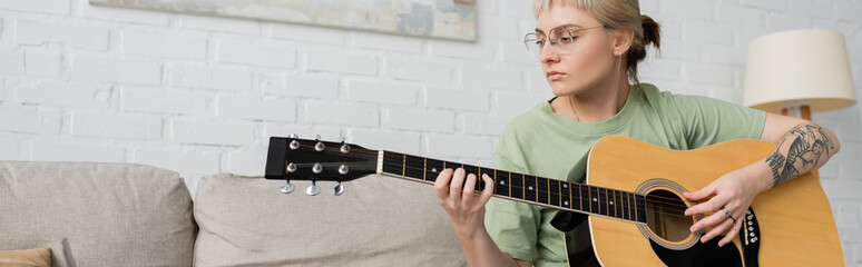 young woman in glasses with bangs and tattoo on hand playing acoustic guitar and sitting on...