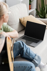 Fototapeta na wymiar young woman in glasses holding acoustic guitar and learning how to play while looking video tutorial on laptop with blank screen and sitting on comfortable couch in living room, guitar lessons