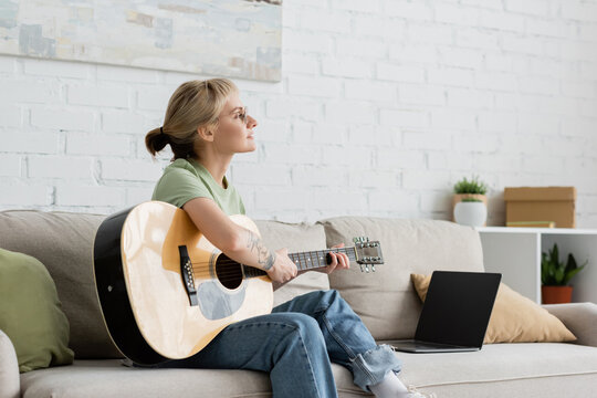 young woman in glasses with bangs and tattoo playing acoustic guitar near laptop with blank screen and sitting on comfortable couch in living room, learning music, skill development