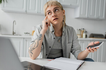 pensive young woman with tattoo on hand and bangs holding smartphone with blank screen and pen near notebook and laptop on white table, blurred foreground, work from home