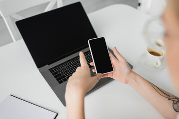 top view of tattooed woman holding smartphone with blank screen near laptop, notebook, and cup of coffee with saucer on white table while working from home, freelancer, modern workspace