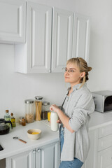 young woman in eyeglasses and short hair with bangs holding bottle with milk near bowl with cornflakes while making breakfast and standing in casual clothes next to kitchen appliances at home