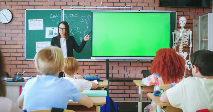 Cheerful Caucasian young male teacher of anatomy or biology bringing scelet to class and explaining human body and bones to pupils. At school. Education concept. Indoors. Teaching.