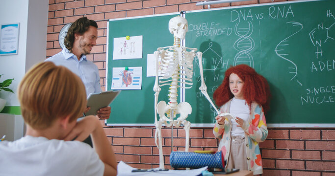 Red haired school girl showing human parts on skeleton while answering questions to male teacher. Biology class at modern school. Education with interest.