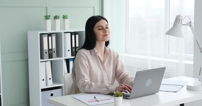A female businesswoman in a shirt is working with a laptop in the office, smiling with a wide grin. She leans back on the chair with a beaming smile, felling joy of successfully completing her work.