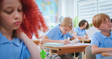 Male and female pupils in blue school uniform sitting at desks white female teacher giving results of control work. Learning process at modern school.