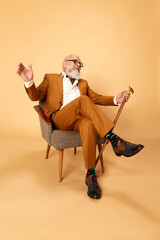 Fototapeta premium Portrait of bearded, mature, bald man in elegant, classical suit and walking stick, sitting in armchair and smiling against studio background. Concept of human emotions, lifestyle, fashion, business