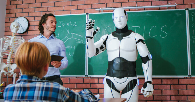 Smart male teacher in blue shirt using digital tablet for controlling modern white robor during interactive class at school. Presentation of robotics during education process.