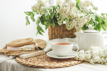 Cup of tea and teapot on windowsill and basket with white lilacs, good morning concept