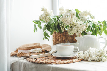Cup of tea and teapot on windowsill and basket with white lilacs, good morning concept