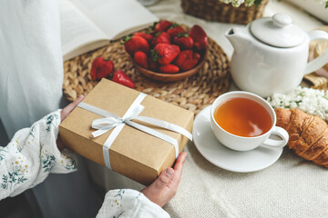 The child's hands hold a beautiful gift box with a ribbon against the backdrop of a festive breakfast. Top view, close-up. Happy mother's day
