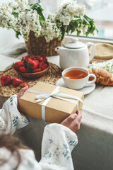 The child's hands hold a beautiful gift box with a ribbon against the backdrop of a festive breakfast. Top view, close-up. Happy mother's day