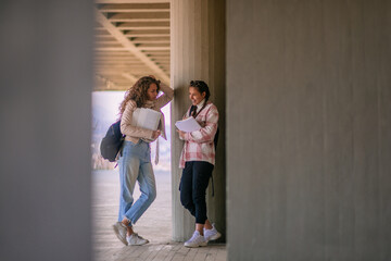 Brunette school girl explaining something to her friend and laughing while leaning on a pillar at the school yard