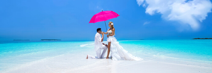 Summer love. Beautiful happy young couple in wedding clothes and pink umbrella is standing on a beach in the Maldives. Engagements and wedding on the beach on the paradise island of Maldives.