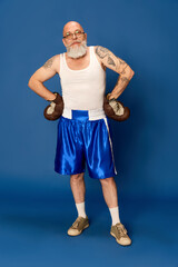 Portrait of bald, bearded, mature man in sportswear and glasses, with tattooed body in vintage boxing gloves against blue studio background. Concept of emotions, sport, active lifestyle, sport fashion