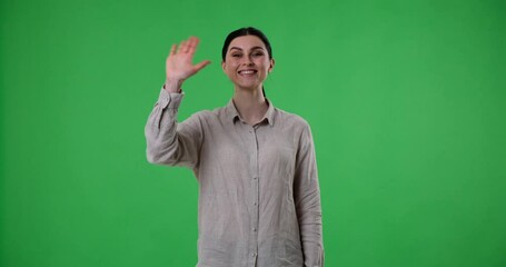 Woman stands on a green screen background, waving her hand in greeting. With a wide smile and a confident expression, she welcomes someone warmly and radiates happiness and joy.
