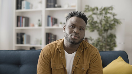 Portrait of a confident and sincere African American man sitting on a sofa at home - obrazy, fototapety, plakaty