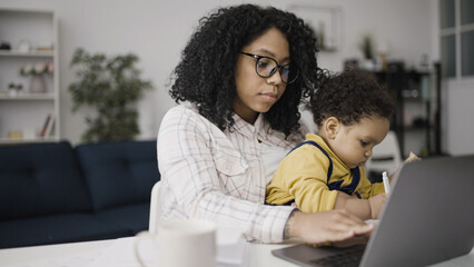 An African American woman is typing on her laptop with her child on her knees drawing