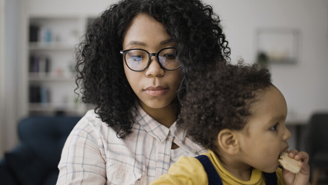 Busy African American woman in eyeglasses working on her laptop with her child sitting in her lap