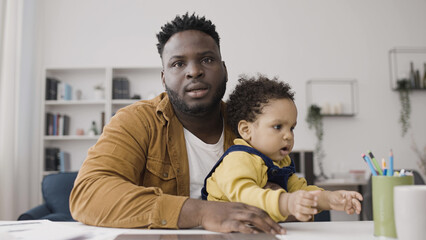 Black father on paternity leave has a video call on his laptop, balancing his work and personal life