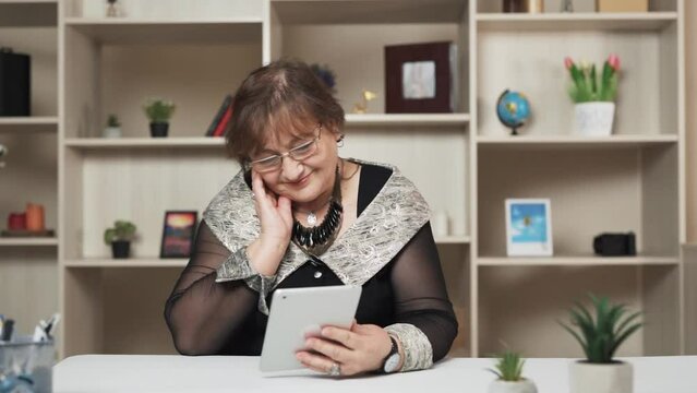 In a modern room, an elder woman is seated at a table, clutching a tablet in her hands. She gazes at its screen, wearing a smile that suggests she is reminiscent of delightful moments from her life.