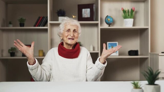 An elderly woman is seated at a table within a room that is both capacious and brightly illuminated. She is passionately relaying information while making numerous gesticulations during her narration.