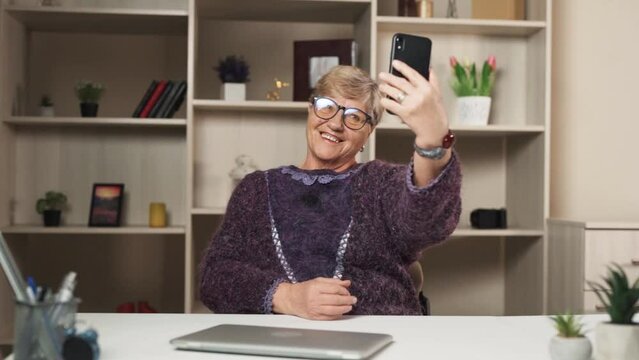 The elderly woman is sitting in a room at the table. There is a computer on the table, and she has a phone in her hands, trying to take a selfie while striking various poses.