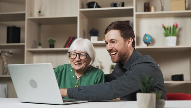 A young man is happily conversing with an elderly woman in a contemporary room while she sits at a computer. He is in the midst of explaining something to her, and both of them wear smiles.