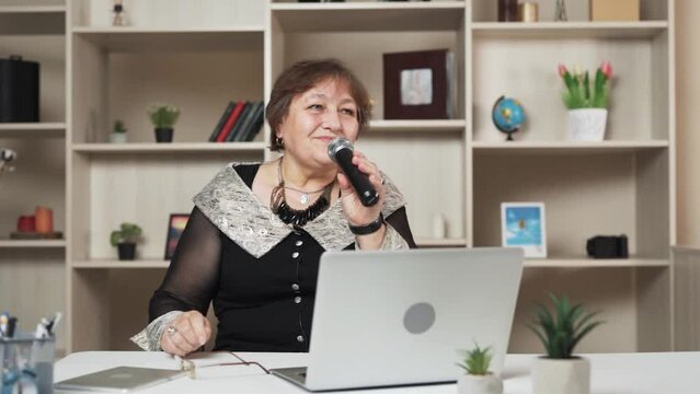 An elderly woman sits in a room with a laptop on her table. She clearly has her webcam turned on and holds a microphone in her hands as she sings. She moves to the rhythm of the music while sitting.