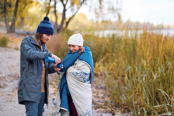 Fototapeta premium Holidays with friend. Young couple drink hot beverage from thermos near river.