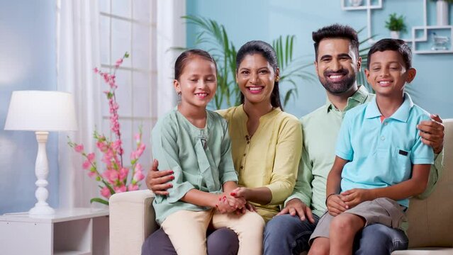 Happy smiling indian couple with sibling kids looking or posing to camera while sitting on sofa at home - concept of nuclaer family, parenthood and generation.