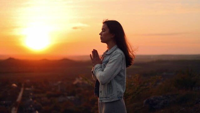 Girl folded hands in prayer silhouette at sunset. woman praying standing outdoors. A woman prays to God, asks for forgiveness of sins and peace in the world