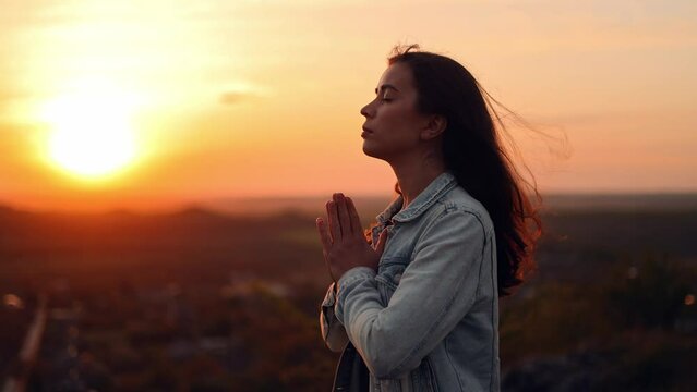 Girl folded hands in prayer silhouette at sunset. woman praying standing outdoors. slow motion video. A woman prays to God, asks for forgiveness of sins and peace in the world