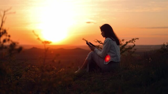 A long-haired girl reads a book in the sun. A young woman reads the Bible outdoors. A woman holds a Bible in her hands and studies the word of God on top of a mountain. Searching for truth