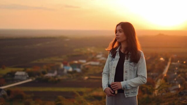 Portrait of a young woman holding a bible in her hands looking at the camera outdoors, smiling reassuringly at sunset. The girl hugs the Bible to her heart